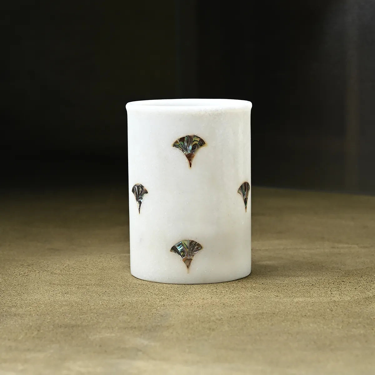 A white cylindrical ceramic cup or candle holder with four small iridescent fan-shaped inlays, set against a dark background on a textured beige surface.